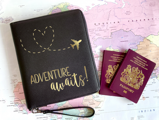 A black faux leather travel wallet for your passports, travel documents, and tickets. Printed with a gold aeroplane and wording Adventure Awaits!