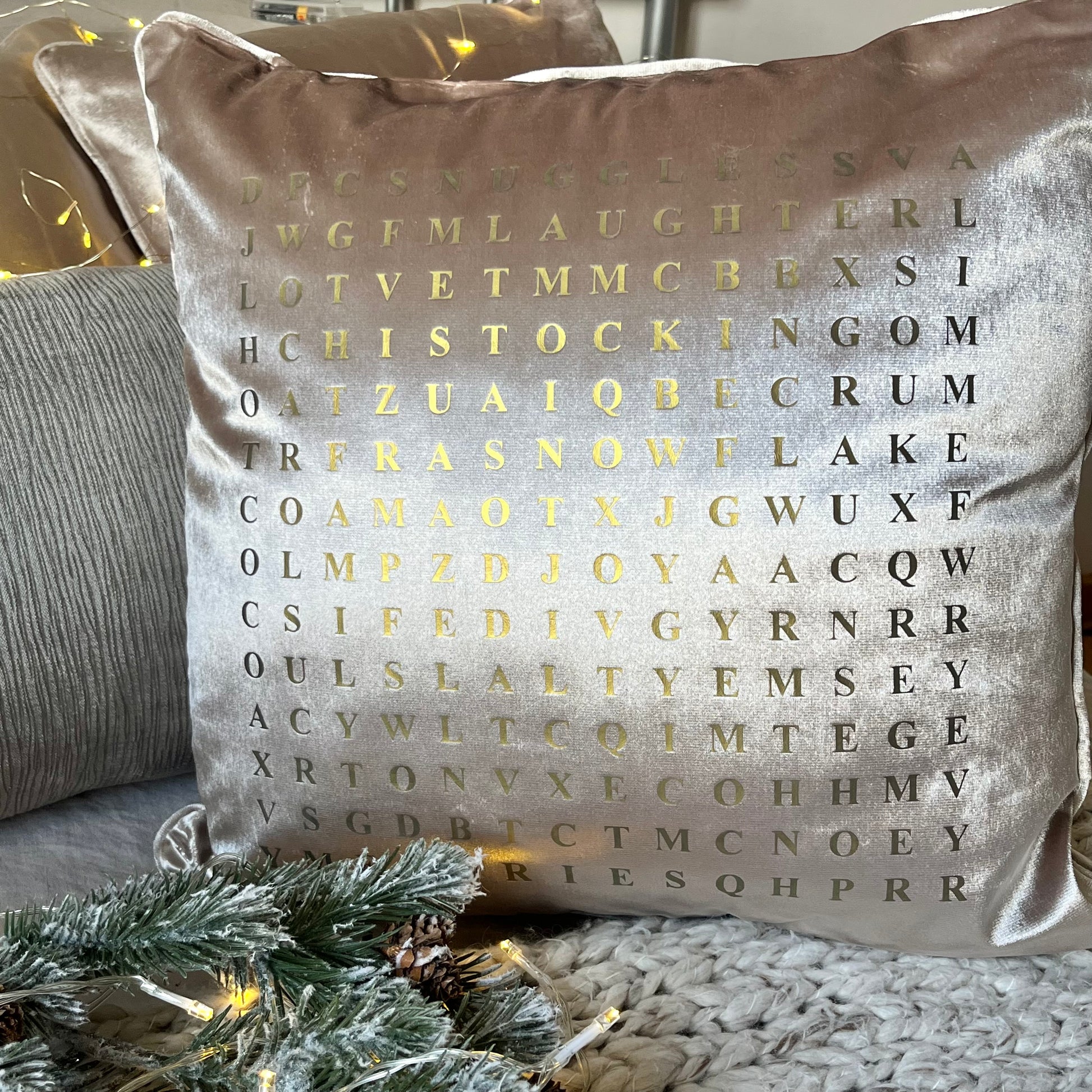 champagne coloured christmas cushion with wordsearch design printed in gold. cushion is placed on a knitted throw with christmas lights behind