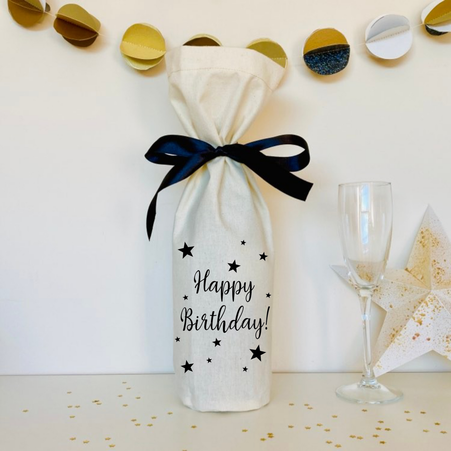 a natural coloured calico gift bag for a bottle of wine. printed with the words Happy Birthday and decorated with stars. The bottle is tied with black ribbon