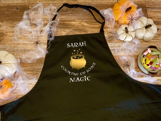 A black apron with a halloween theme, a witches cauldron with the wording 'Cooking up some Magic', personalised with a name